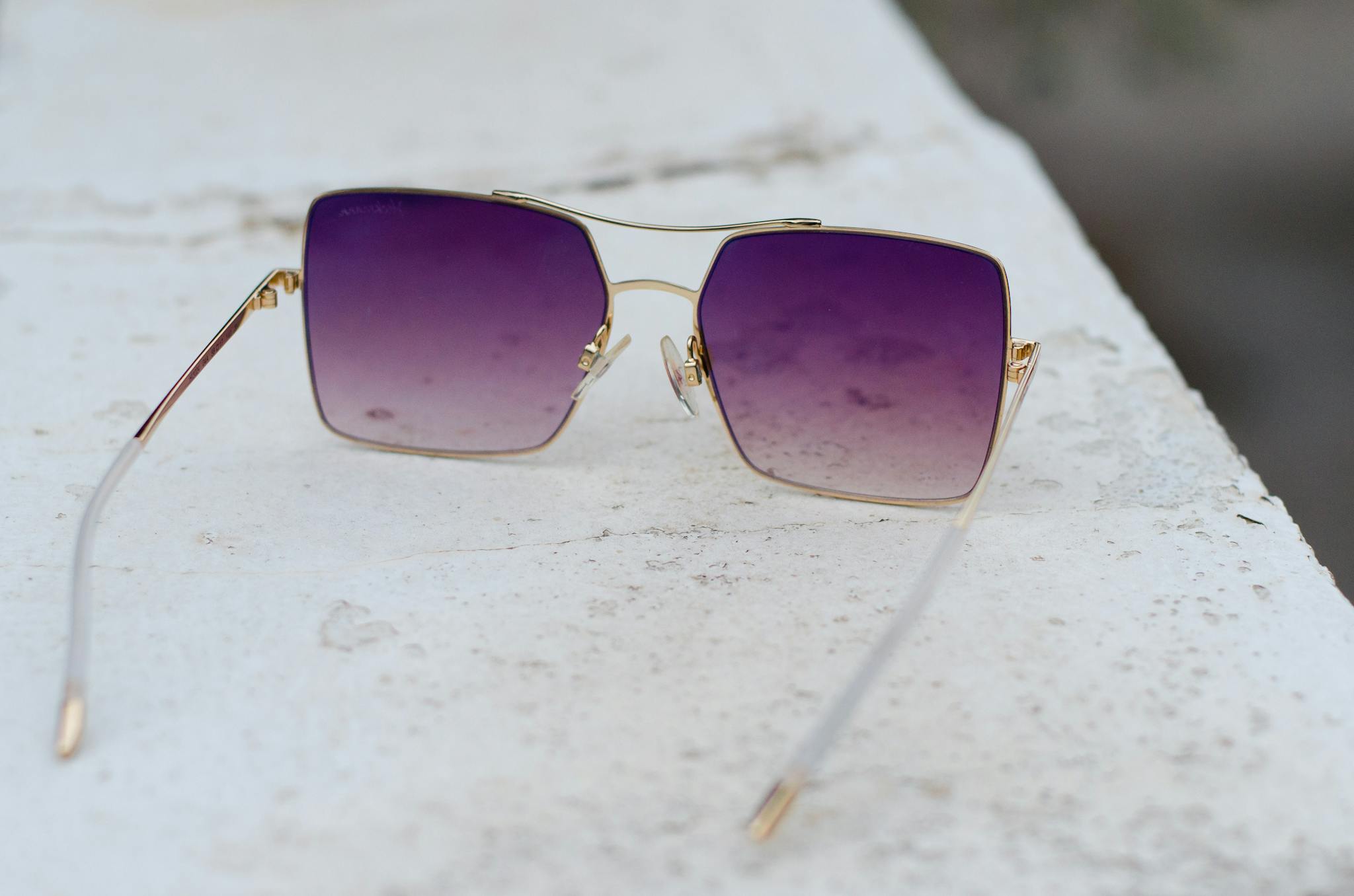 Close-up of stylish square sunglasses with purple gradient lenses on a textured white surface.